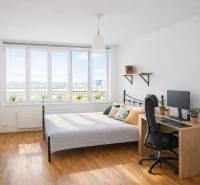 Ložnice v dvoupokojovém bytě s postelí, stolem, počítačem a podlahou s dřevěným dekorem.