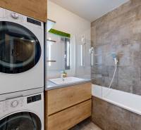 Koupelna v 3-pokojovém bytě s pračkou, sušičkou, vanou a šedými obklady.