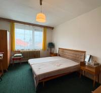 Ložnice s jednoduchým nábytkem v rodinném domě, zelený koberec a žluté záclony.