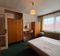 Ložnice rodinného domu s kobercovou podlahou, dřevěným nábytkem a velkým oknem.