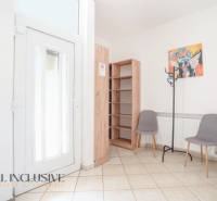 Interiér budovy se vstupními dveřmi, dlažbou, poličkou, dvěma židlemi a věšákem.