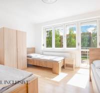 Světlý pokoj se třemi postelemi, bílými stěnami, skříněmi a velkými okny.