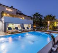 Vila v Kaštela s osvětleným bazénem, lehátky a palmami v zahradě při soumraku.