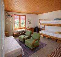 Pokoj v chatě s palandami, zelenými křesly a velkým oknem.