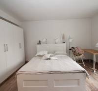 Ložnice v jednopokojovém bytě s postelí, bílou skříní a podlahou s dřevěným dekorem.