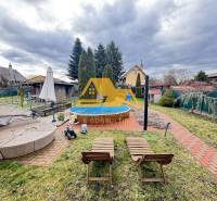 Zahrada u chaty s bazénem na ulici v Nových Zámcích, obklopená udržovanou zelení.