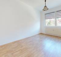 Pokoj v 3-pokojovém bytě s bílými stěnami, podlahou s dřevěným dekorem a oknem.