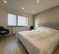 Ložnice v dvoupokojovém bytě s podlahou s dřevěným dekorem, pohodlným křeslem a velkým oknem.