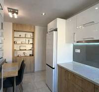 Kuchyně v 2-pokojovém bytě s bílými skříňkami, stolem a dřevěným dekorem polic.