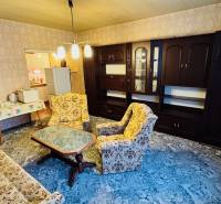 Obývací pokoj v dvoupokojovém bytě se starším nábytkem a kobercem na podlaze.