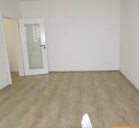 Pokoj v 3-pokojovém bytě s bílými stěnami a podlahou s dřevěným dekorem.