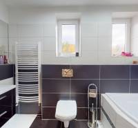 Koupelna v rodinném domě s vanou, dlaždicemi, umyvadlem a okny na stěně.