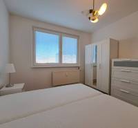 Ložnice v dvoupokojovém bytě s bílým nábytkem a postelí, okno se žaluziemi.