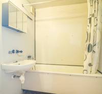 Koupelna s vanou a sprchovým závěsem v 2-pokojovém bytě, světlé stěny, oranžová podlaha.