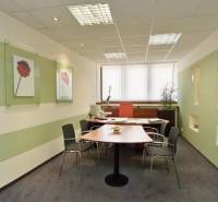 Kanceláře s pracovním stolem, židlemi, umístěné dekorace na stěnách a okno s vertikálními žaluziemi.