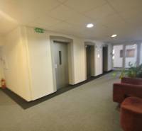 Interiér kanceláře s výtahy a sedačkou, bílá stěna, piktogramy “EXIT”.