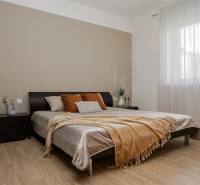 Ložnice v dvoupokojovém bytě s podlahou s dřevěným dekorem, velkou postelí a oknem s jemným závěsem.