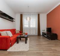 Obývací pokoj s oranžovou sedačkou, poličkami a podlahou s dřevěným dekorem v 2-pokojovém bytě.