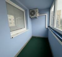 Zasklená lodžie s klimatizační jednotkou v 2-pokojovém bytě, vybavená žaluziemi a zeleným kobercem.