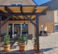 Gastro prostory v Nové Dědince s pergolou, květináči a venkovním posezením na dlažbě.