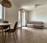 Garsonka na ulici Bodona s dřevěným nábytkem, gaučem, stolem, balkónem a kuchyňskou linkou.