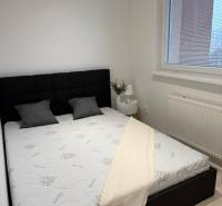 Ložnice v dvoupokojovém bytě s černým čelem postele, bílou přikrývkou a oknem s roletami.