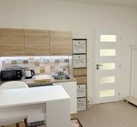 Garsonka se světlou kuchyňskou linkou, bílým barovým pultem a podlahou s dřevěným dekorem.