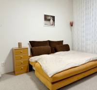 Garsonka s postelí, nočním stolkem a kobercem, jemné závěsy na okně.