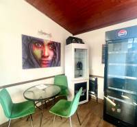 Gastro prostory s podlahou s dřevěným dekorem, zelené židle, obrazy a lednička.