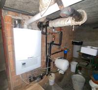 Plynový kotel ve sklepě rodinného domu s potrubím a zásobníkem vody na stěně.