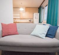 Šedá pohovka s barevnými polštáři v 4pokojovém bytě u okna a kuchyně.