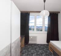 Pokoj ve 4pokojovém bytě s postelí, tmavými závěsy a výhledem z okna.