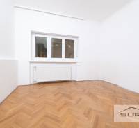 Pokoj ve 4pokojovém bytě s bílými stěnami a podlahou s dřevěným dekorem.