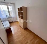 2-pokojový byt, postel u okna, regál, podlaha s dřevěným dekorem, bílá stěna.