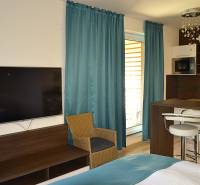 Garsonka s TV, dřevěným nábytkem, kuchyňským pultem a tyrkysovými závěsy.