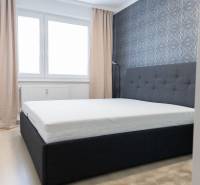 Ložnice ve 4pokojovém bytě s postelí, tapetou, okny a závěsy, podlaha s dřevěným dekorem.