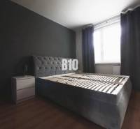 Ložnice s tmavě šedou postelí, nočním stolkem a podlahou s dřevěným dekorem ve 3pokojovém bytě.