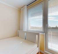 Postel v garsonce s výhledem přes žaluzie na okně.