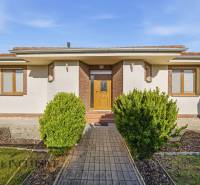 Rodinný dům v Kútnikách s keřovou výsadbou, bílou fasádou a hnědými doplňky.