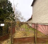 Starý plot brání vstupu na zarostlý pozemek. Rodinný dům s bílou fasádou na boku.