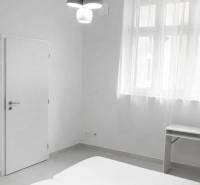 Bílý pokoj s manželskou postelí, stolem a závěsným osvětlením v 2pokojovém bytě.