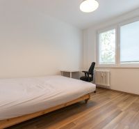 Ložnice s jednoduchou postelí, pracovním stolem a podlahou s dřevěným dekorem ve 3-pokojovém bytě.