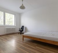 Světlá ložnice s postelí a pracovní židlí, podlaha s dřevěným dekorem.