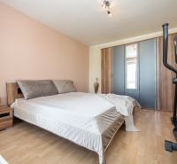 Ložnice v 2pokojovém bytě s postelí, skříní, cvičebním strojem a podlahou s dřevěným dekorem.