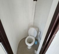 Jednoduchá toaleta v chatě se stěnou bez obkladu a plastovým WC.