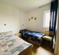 Dvě postele, židle, okno, obrazy na stěnách, podlaha s dřevěným dekorem.
