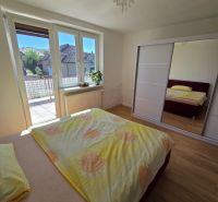 Ložnice v rodinném domě s postelí, skříní, oknem na balkon a podlahou s dřevěným dekorem.