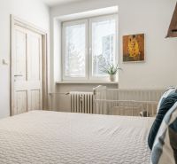 Ložnice v třípokojovém bytě s postelí, rostlinou a dekorativním obrazem na stěně.