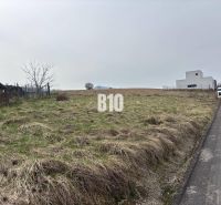 Pozemek na bydlení v Rosině, vedle cesty, s travnatým povrchem a nedalekými stavbami.