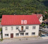 Budova v Belej s červenou střechou, obklopená zelení a cestou v popředí.